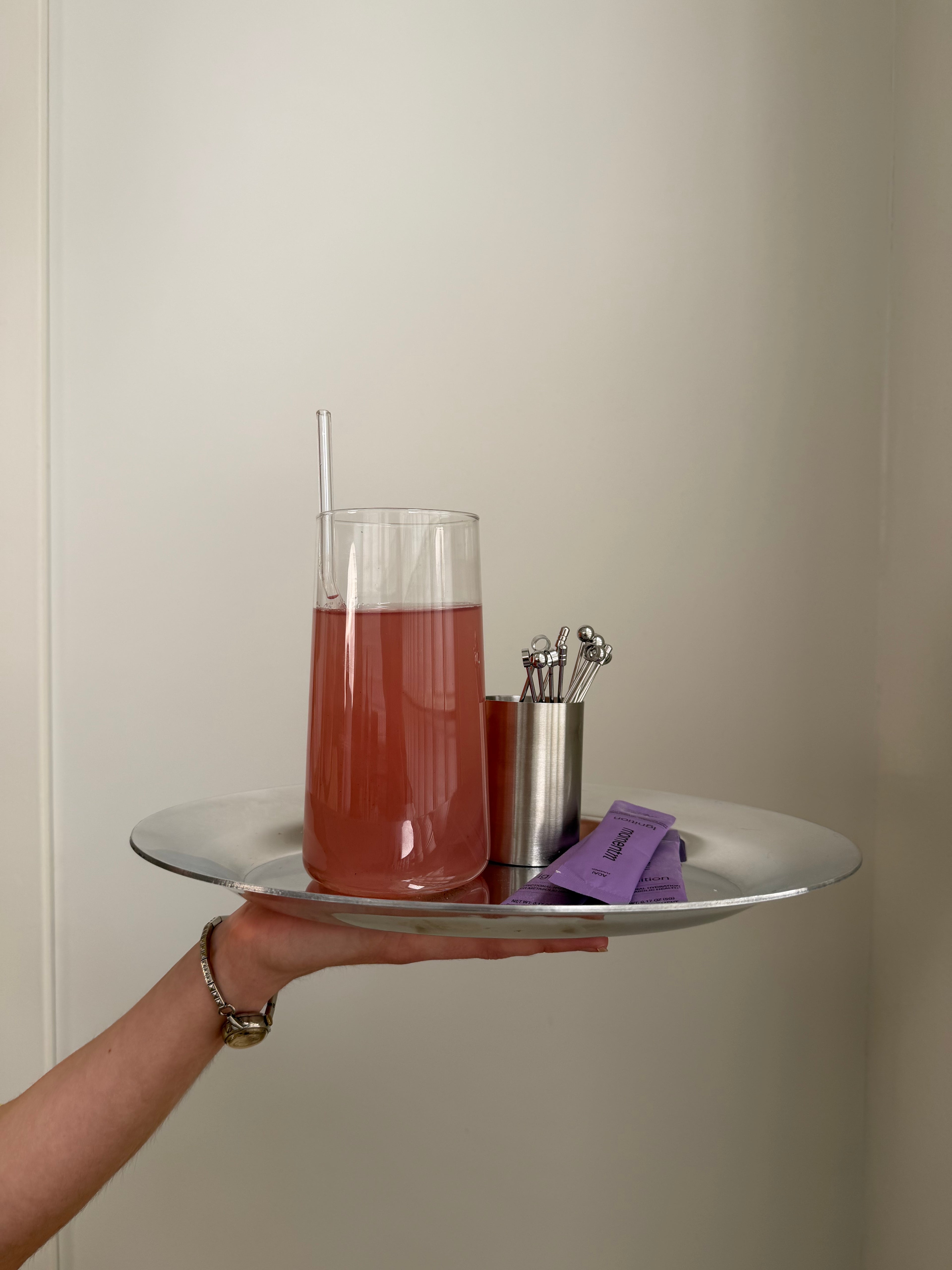 Hand holding a tray with a glass of red liquid, straws, and a small container against a plain background