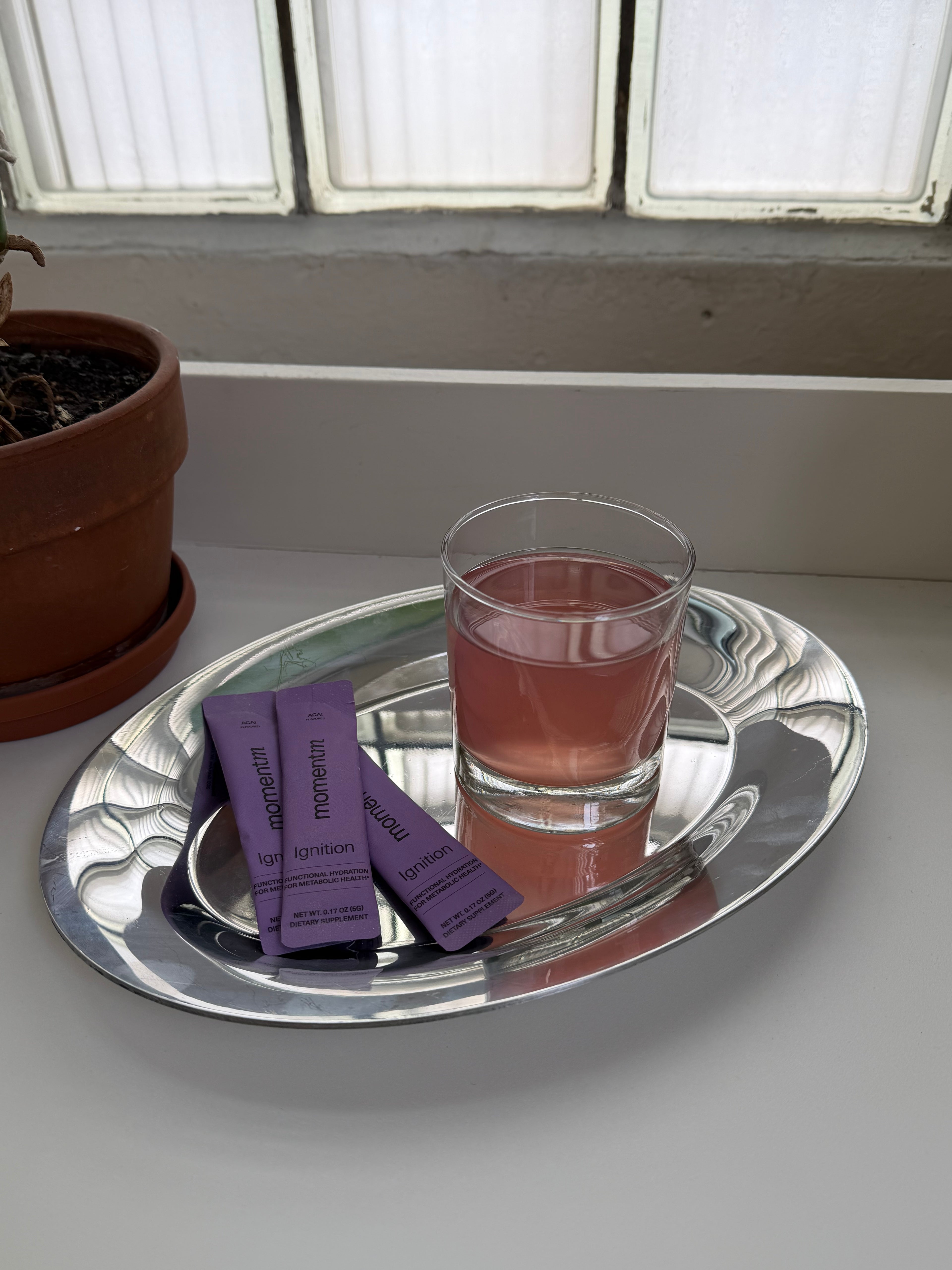 Glass of pink liquid and purple packets on a silver tray with a window in the background