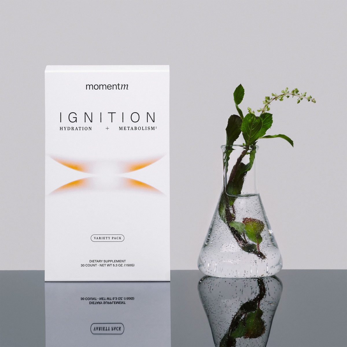 momentén Ignition supplement box with a clear vase containing green leaves on a reflective surface.